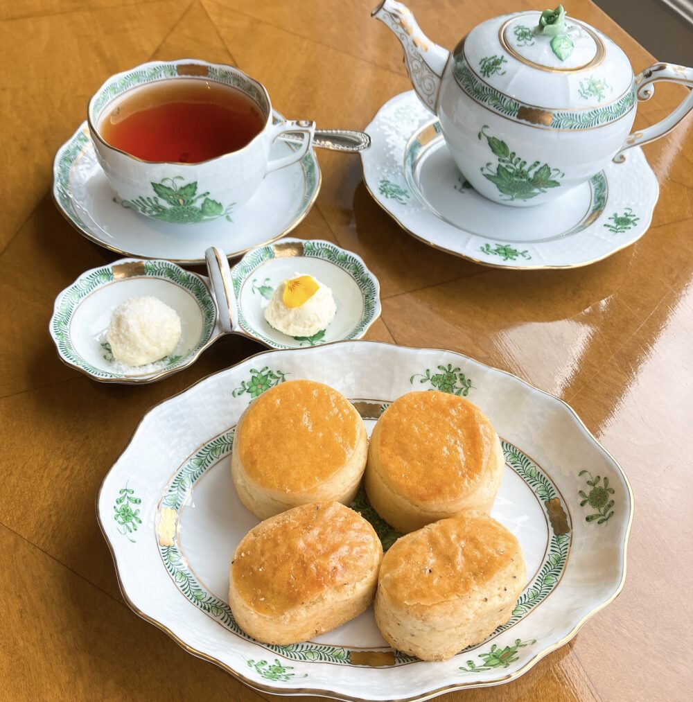 木目のテーブルに置かれた、英国女王も愛した洋食器ブランド「ヘレンド」のティーセット。手前にはプレーンとトリュフの2種類のスコーンが盛られた皿、奥には紅茶の入ったカップ＆ソーサーとティーポット、クロテッドクリームとジャムを乗せる小皿。