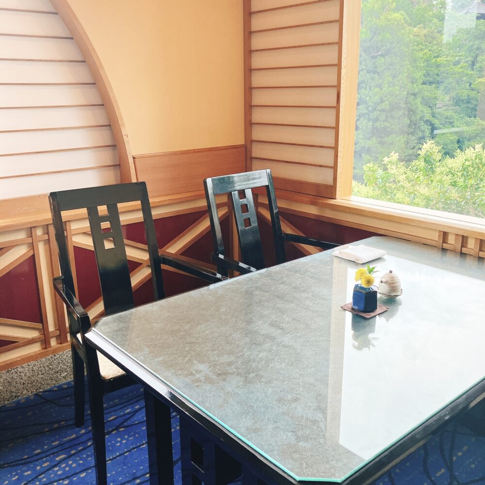 日本庭園が見える窓際に配置された、日本料理レストラン「みゆき」のテーブル席。木製の椅子が二脚とガラス天板のテーブルが手前にあり、窓の外には緑豊かな木々が広がる。