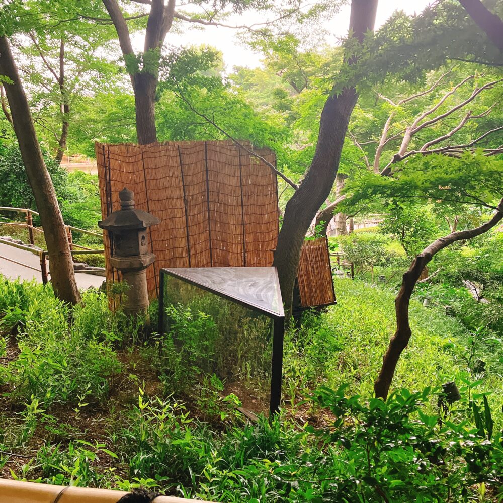 ホテル椿山荘東京の日本庭園内にある、竹の柵と石灯籠が立つ草木の茂る一角。手前には三角錐のオブジェのようなものが設置されており、奥は木々に覆われている
