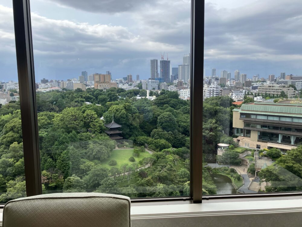 ホテル客室の窓から広がる、緑豊かな日本庭園。園内には三重塔が見え、奥には都会のビル群が広がる。