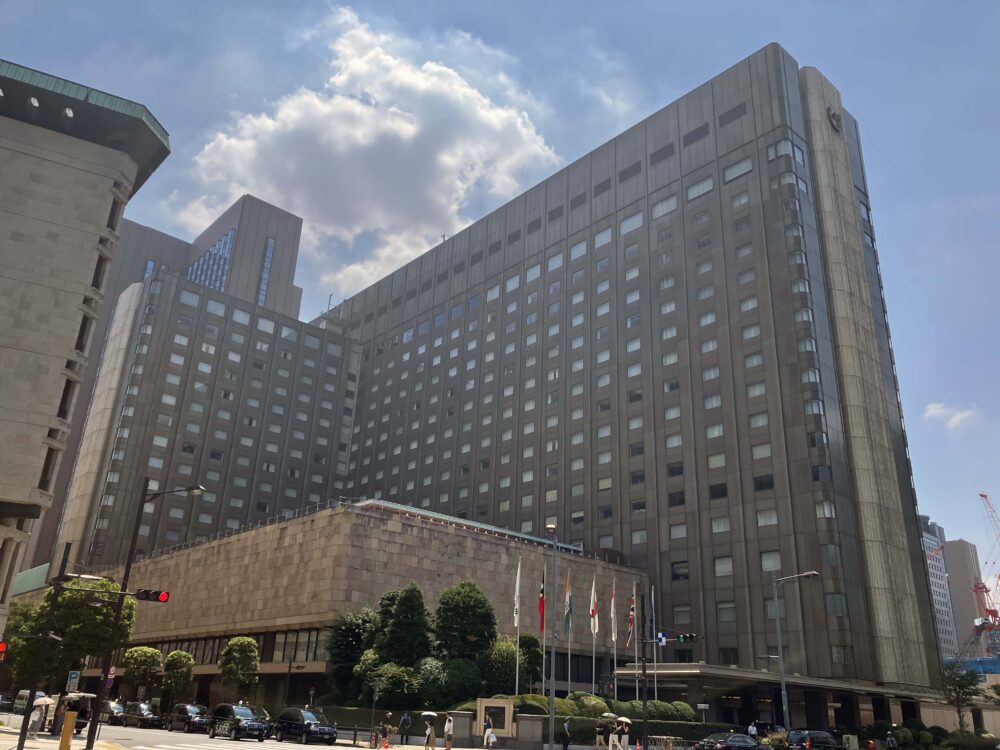 2025年8月撮影の帝国ホテル東京本館(3代目建物)の外観。青空の下、重厚なグレーのファサードが特徴的な建物がそびえ立ち、敷地前には数本の旗が掲げられている。