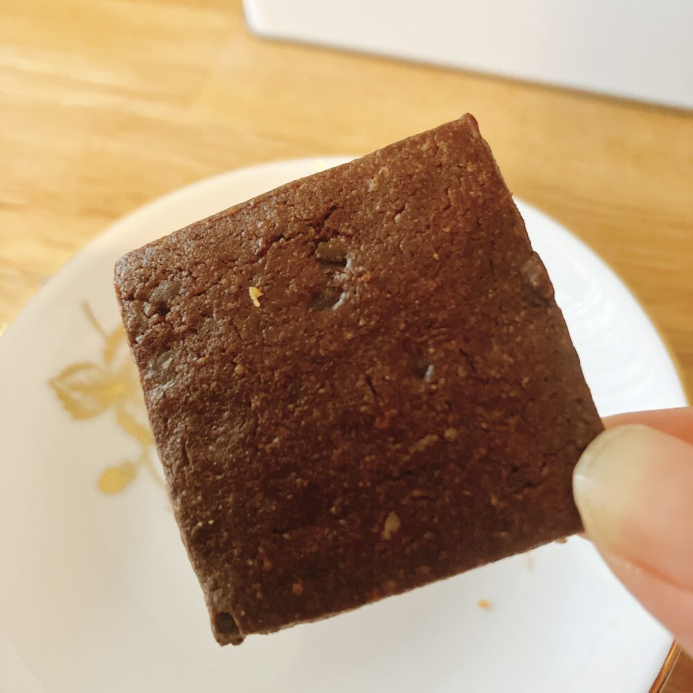 帝国ホテルのプレミアムクッキー缶に入っている、手で持たれた濃い茶色の四角いサブレショコラ。カカオの深みを感じる濃厚なチョコレートクッキー。