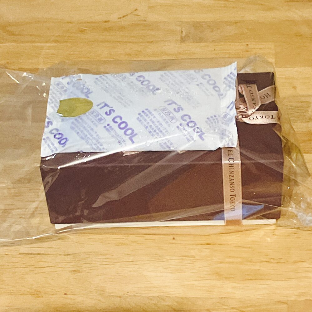 ホテル椿山荘東京の栗のパウンドケーキの箱(濃い茶色)が、透明なビニール袋に入れられ、上に「IT'S COOL」と書かれた大きな保冷剤が乗せられている様子。