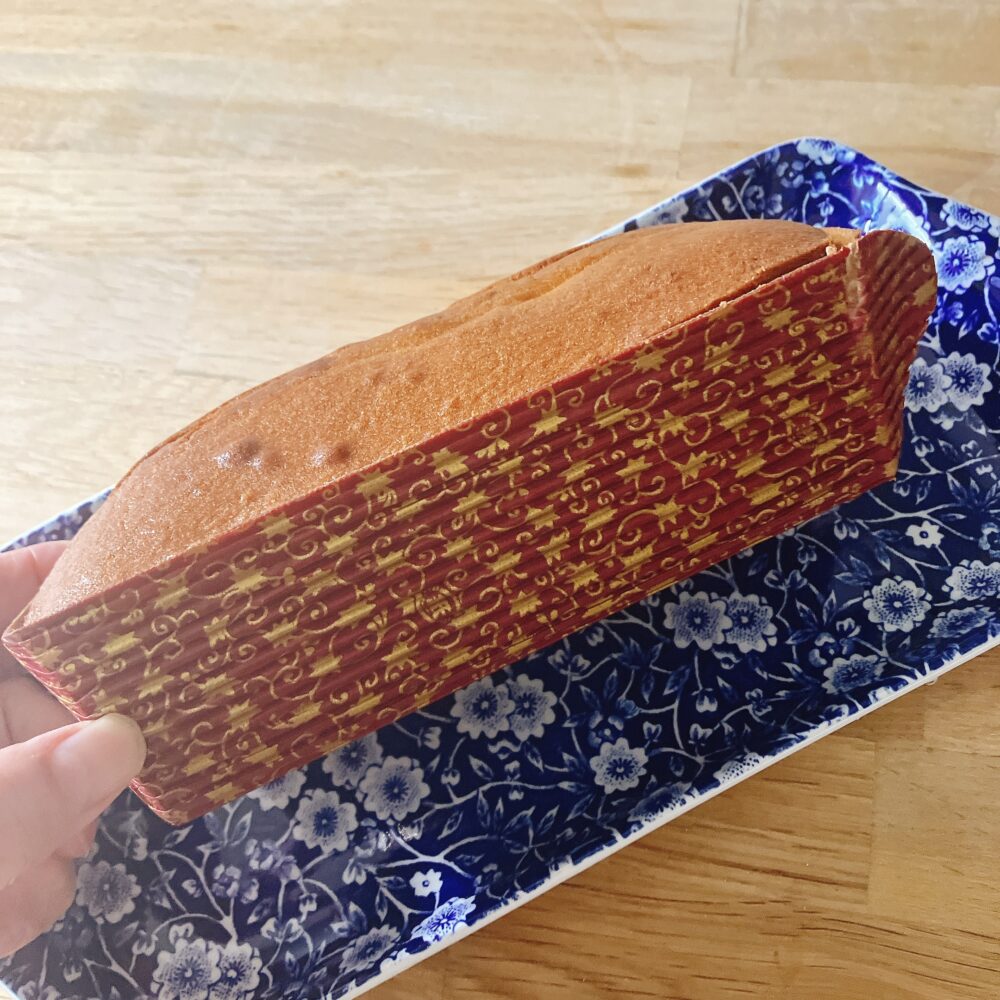 ホテル椿山荘東京の栗のパウンドケーキが、赤と金色の華やかな柄の型紙に包まれた状態で、青と白の模様の皿の上に置かれている。右上の部分が少し剥がされている。
パウンドケーキは、取り出しやすいように型紙に包まれた状態。赤と金の華やかなデザインで、このままテーブルに出しても絵になります。