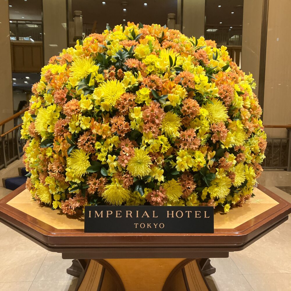 帝国ホテル東京本館ロビー中央に飾られた、黄色とオレンジ色の菊を基調とした球状の巨大なフラワーアレンジメント。その手前には「IMPERIAL HOTEL TOKYO」のロゴサインが置かれている。