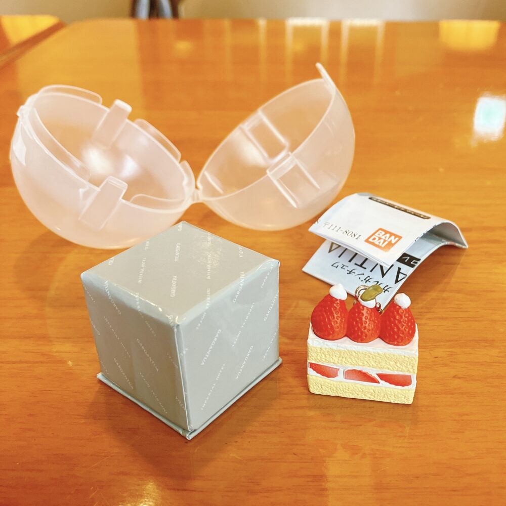 帝国ホテル ガルガンチュワのガチャポン景品。開いたプラスチックカプセルと、景品のストロベリーショートケーキのミニチュア、グレーの箱、説明書が写っている。