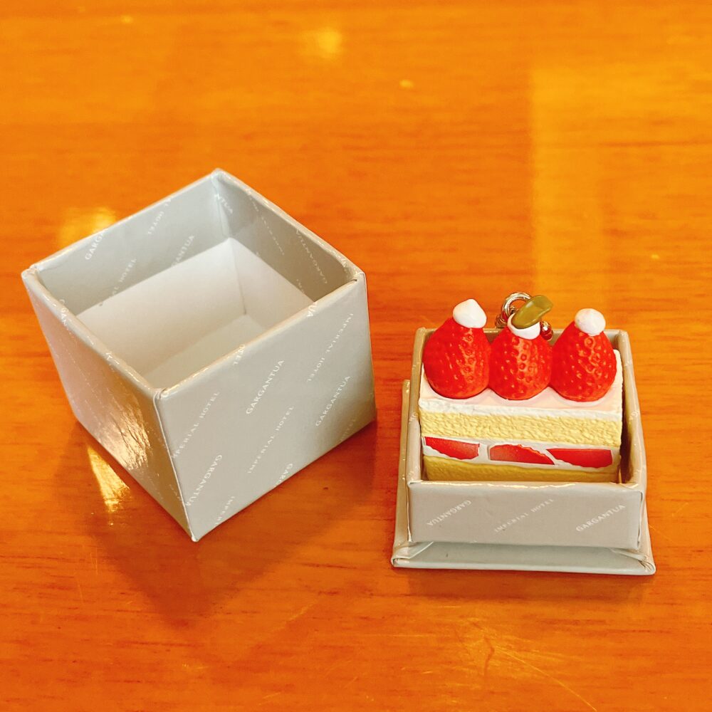 帝国ホテル ガルガンチュワのガチャポン景品。ストロベリーショートケーキのミニチュアが、ガルガンチュワのロゴが入った小さなグレーの紙箱にきれいに収まっている。