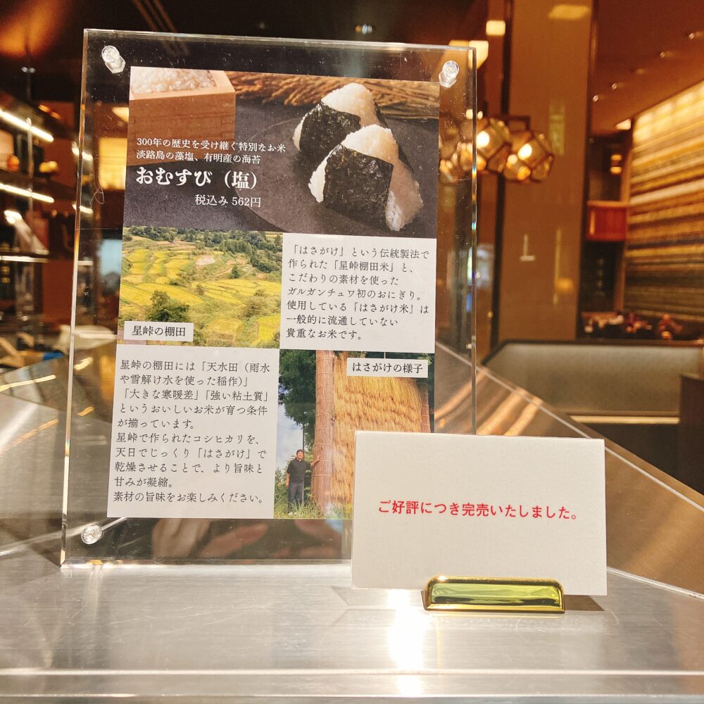 帝国ホテルのガルガンチュワに設置された、「おむすび（塩）」の商品説明ボード。価格は税込562円で、写真の右下には「ご好評につき完売いたしました」という赤い札が置かれている。