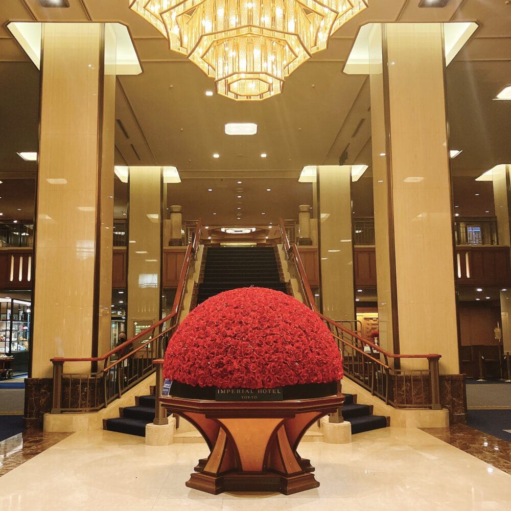帝国ホテル本館メインロビーの象徴である「赤いバラのドーム状の装花（インペリアルレッド）」。背後には重厚な階段と輝くシャンデリアが見える、伝統と格式を感じさせる内観写真。