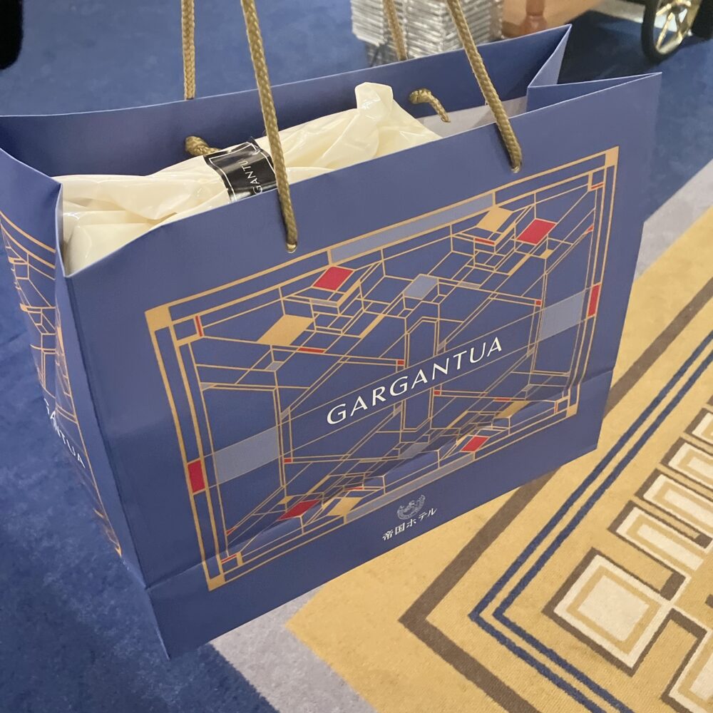 帝国ホテル ガルガンチュワの青い専用ショッパー。アール・デコ調の金色と赤で幾何学模様がデザインされており、バッグの口からは商品の白い包装が見える。