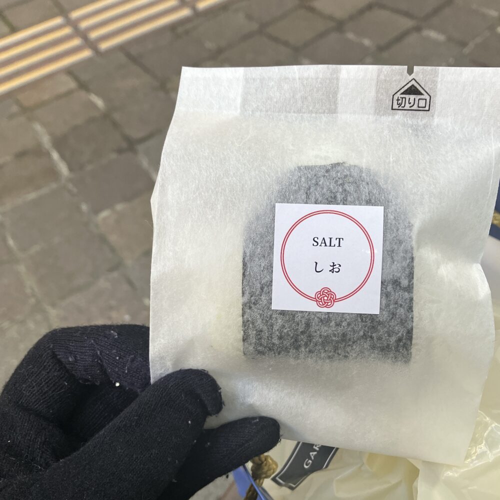 帝国ホテルのおむすび（塩）の個別包装を手に持った写真。白い和紙のような袋に「SALT しお」と日本語と英語で書かれた円形の赤いラベルが貼られ、水引のような結び目のデザインが施されている。