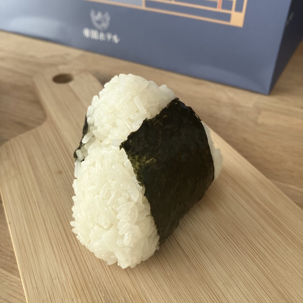 帝国ホテルの「おむすび（塩）」のクローズアップ写真。竹製まな板の上に乗せられており、米粒が一粒一粒しっかりと立っていて、ふっくらと握られている。