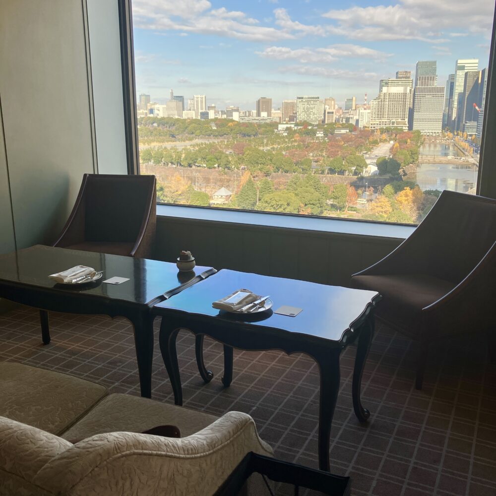 インペリアルラウンジ アクアの窓際席。大きな窓からは秋色に色づいた日比谷公園の木々と皇居の石垣、東京のビル群が一望できる。テーブルには二人分のカトラリーが整然とセットされている。