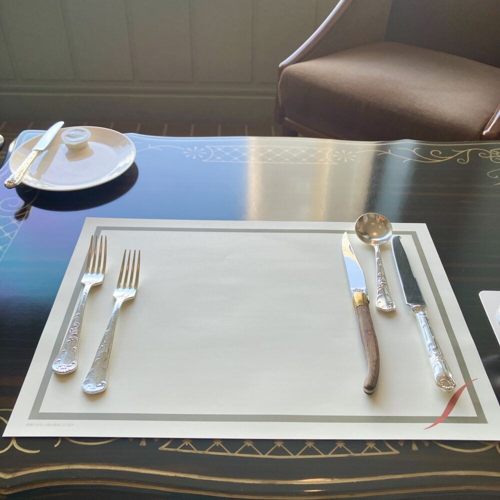 帝国ホテルのロゴが入ったランチョンマットの上に、美しく磨き上げられた銀のカトラリー（ナイフ、フォーク、スプーン）が整然と並べられている。左上にはバターとパン皿が配置された、フルコース開始前のテーブルセッティング。