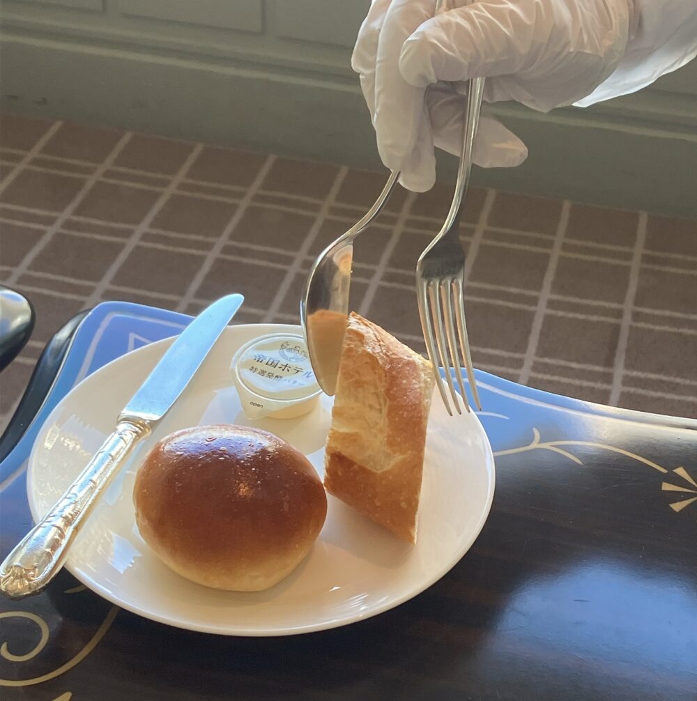 帝国ホテルのスタッフが白い手袋を着用し、フォークとスプーンを片手で操って（ゲリドン・サービス）、白い皿の上にバゲットを取り分けている様子。皿の上には丸いテーブルロールと「帝国ホテル特選発酵バター」が置かれている。