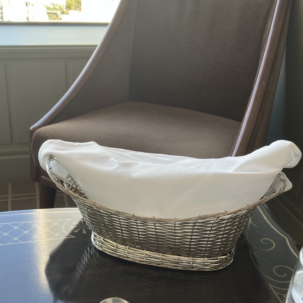 帝国ホテル「アクア」のテーブルに置かれた、白い布でパンを包んだ銀のバスケット。背景には茶色のラウンジチェアが見える。