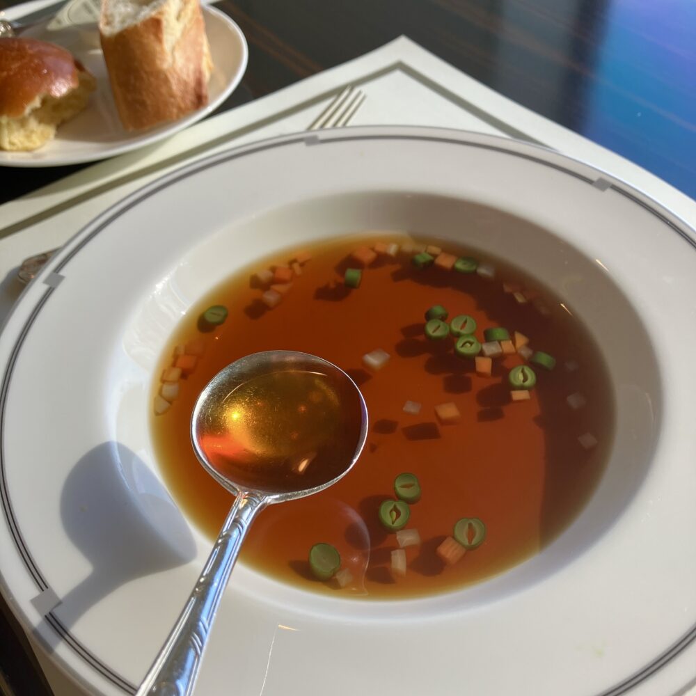 帝国ホテル「インペリアルラウンジ アクア」のダブルビーフコンソメ。スプーンに掬われたスープが琥珀色に透き通り、背後には伝統のテーブルロールとバゲットが並んでいる。