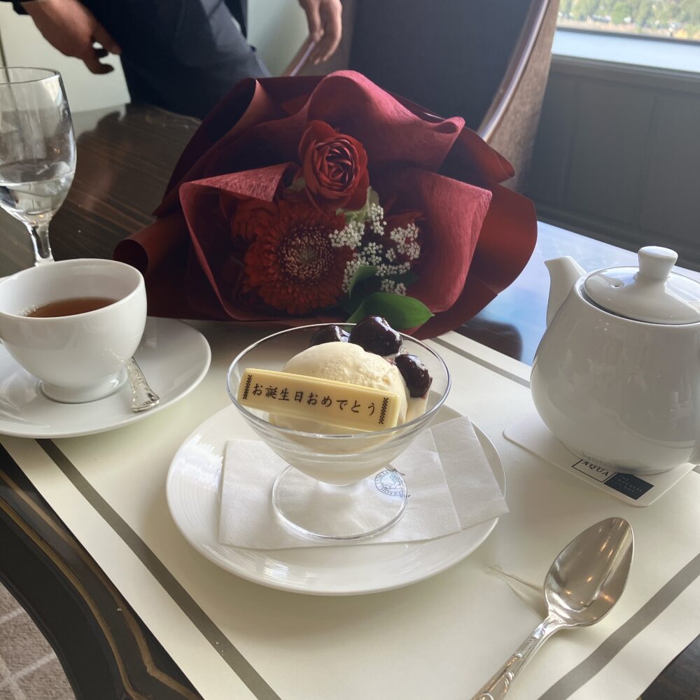 帝国ホテルのレストランで提供された、お誕生日祝いの花束とデザートプレート。深紅のバラ4本と大輪のガーベラ、カスミソウをあしらった気品ある赤いブーケが、バースデープレートの横に添えられている。

バニラアイスに温かいチェリーソースがかかった伝統のデザート「チェリージュビリー」。手前には「お誕生日おめでとう」のチョコプレートが乗っており、奥にはお祝いの花束と白いティーポットが見える。