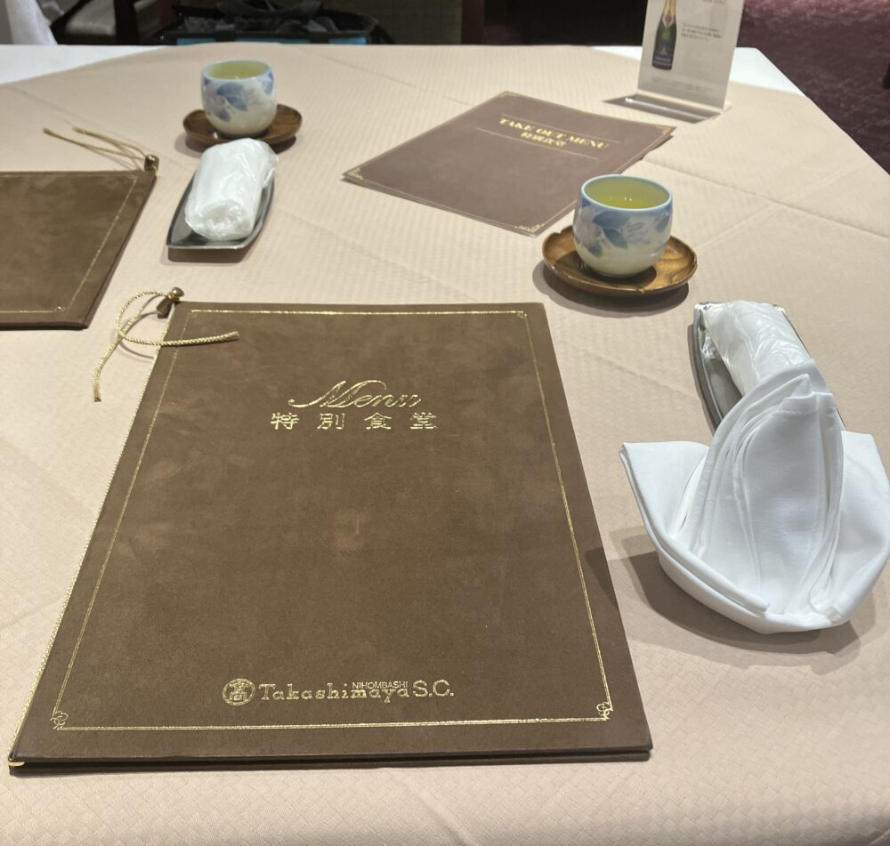 テーブルセッティングの様子。スエード調の重厚なメニューブックの横には、洋食用の白いナプキンと、日本茶（煎茶）の湯呑みが並んでいる。和洋が混在する独特のスタイル。