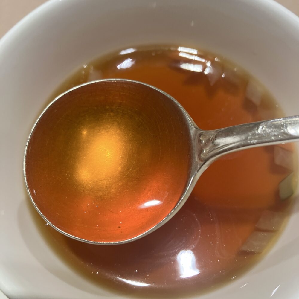 銀のスプーンですくい上げられたダブルビーフコンソメスープのクローズアップ写真。一切の濁りや不純物がない完璧な透明度と、深く輝く琥珀色が、帝国ホテルの卓越した技術を物語っている。