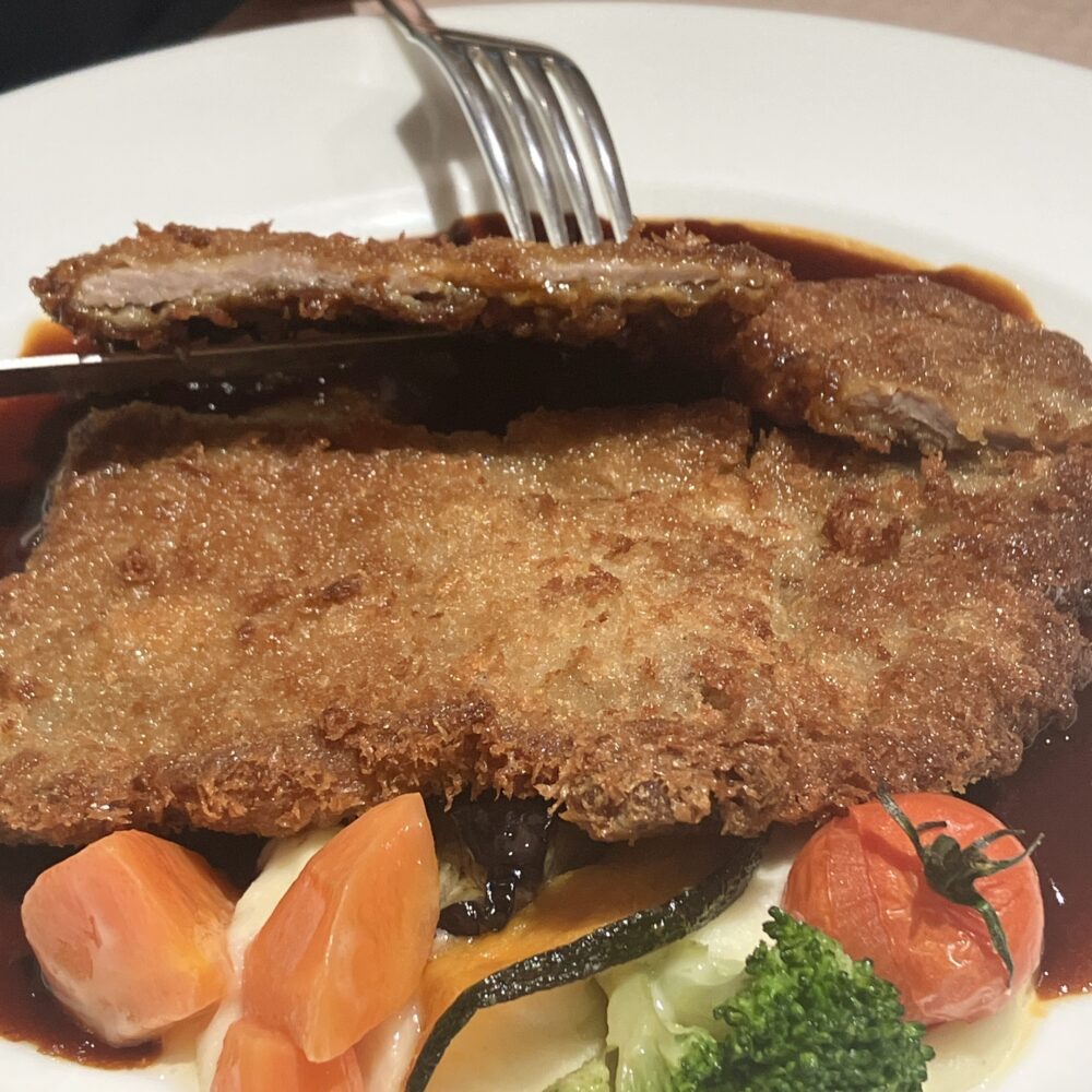 ナイフを入れた仔牛のカツレツの断面。薄く叩いて伸ばされた柔らかい仔牛の肉と、香ばしく揚げられた薄い衣、そしてたっぷり敷かれたデミグラスソースの層が確認できる。