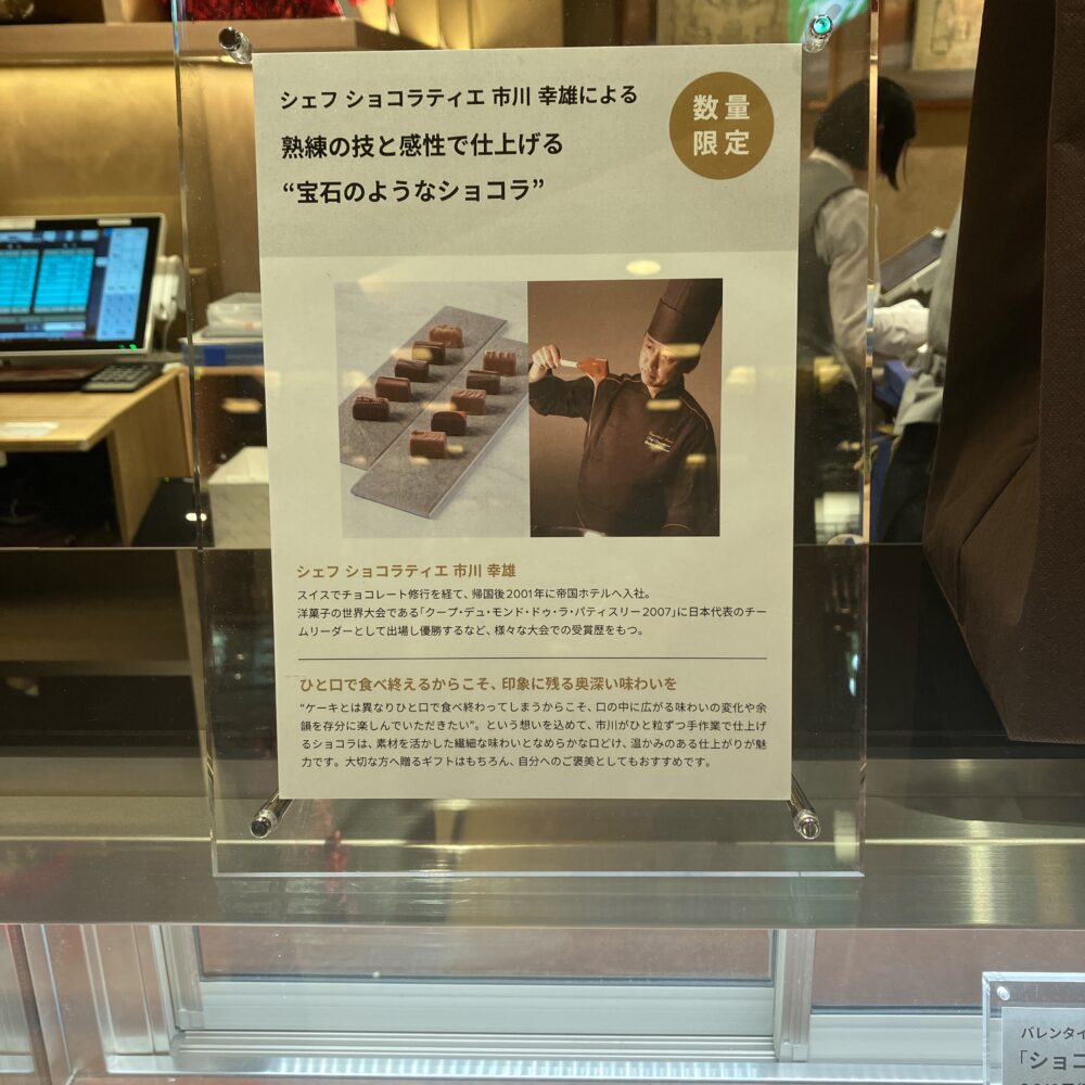 「シェフ ショコラティエ 市川幸雄による熟練の技と感性で仕上げる“宝石のようなショコラ”」と題された店頭パネル。市川シェフがショコラを作っている写真と、クープ・デュ・モンド優勝などの経歴が記されている。