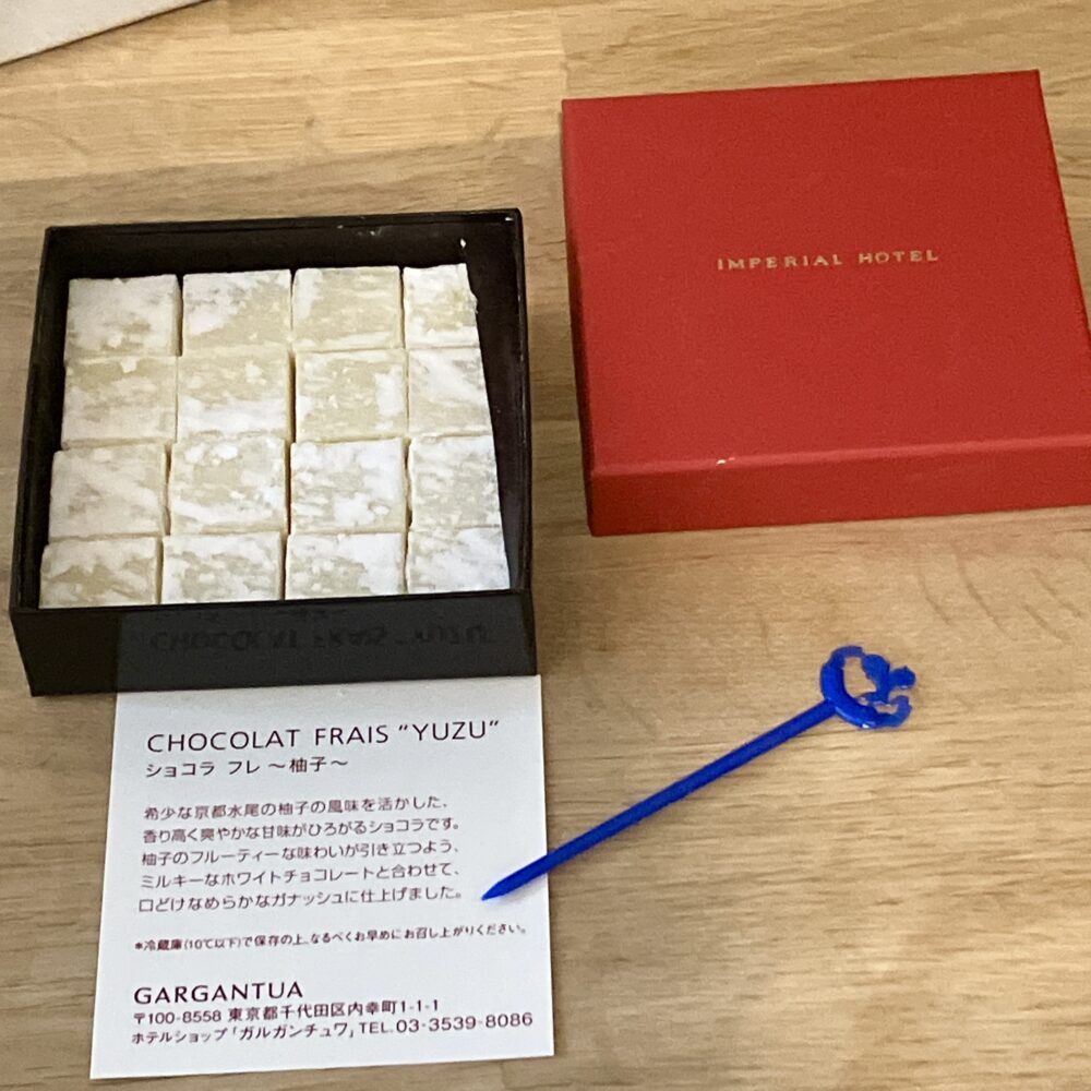 鮮やかな赤の箱と、整然と並ぶ16粒の「ショコラ フレ ～柚子～」。手前には青い帝国ホテルのロゴ入りピックと、京都水尾産の柚子を使用したことを説明するカードが添えられている。