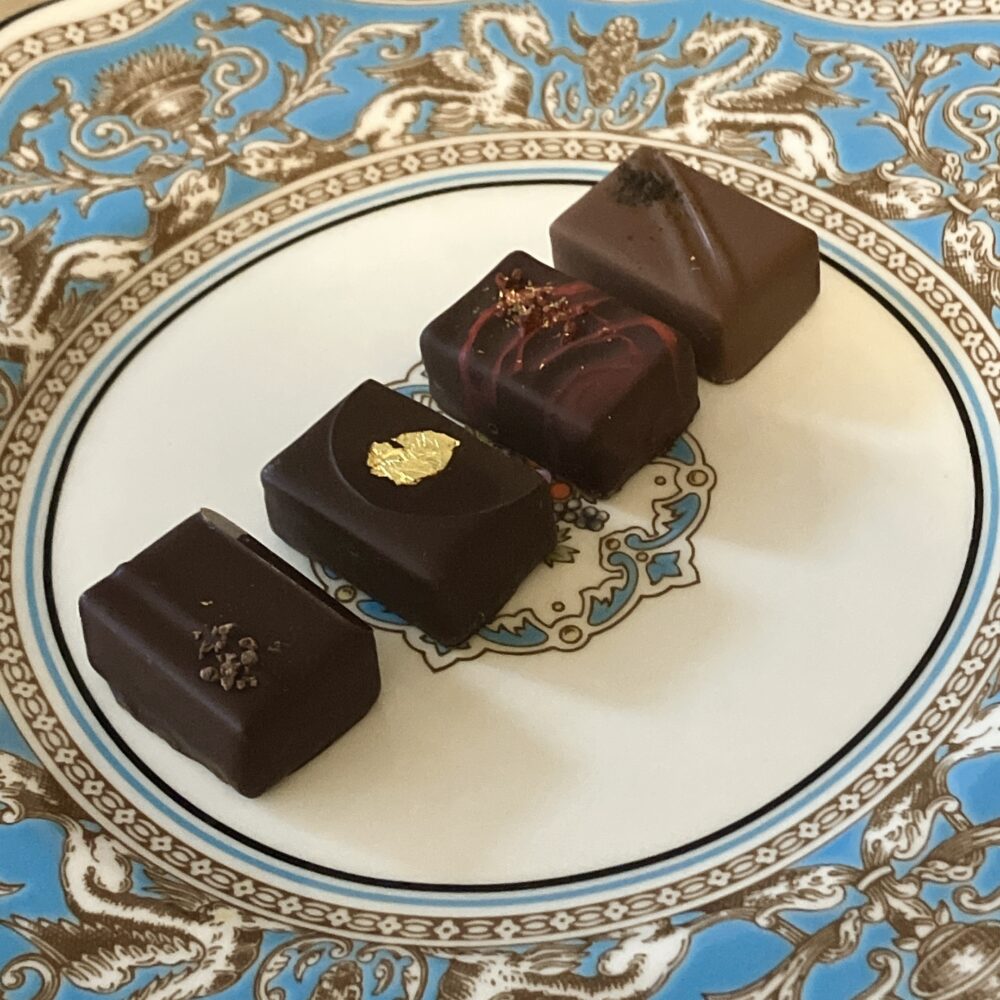 ウェッジウッド「フロレンティーン ターコイズ」の皿に並べられた4粒のボンボンショコラ。手前から奥へ、カカオニブが乗った「ショコラ」、金箔の「カネル」、赤いラインの「フランボワーズ」、茶色のラインの「テ ノワール」が美しく配置されている。