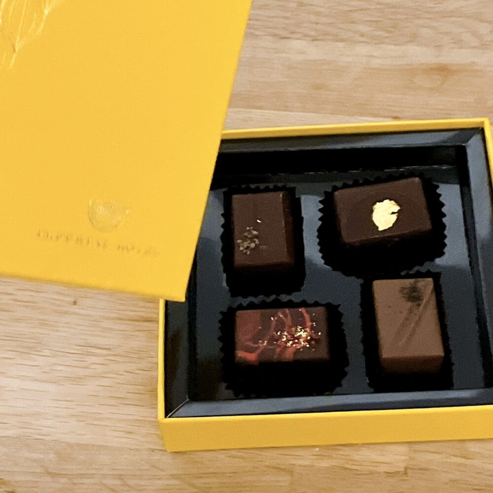 鮮やかな黄色のパッケージに入った「ボンボン ショコラ バレンタインセレクション 2026（4個入）」。蓋を開けた状態で、金箔があしらわれた「カネル」、赤いラインの「フランボワーズ」、カカオニブが乗った「ショコラ」、シンプルな「テ ノワール」の4粒が高級感のある黒いトレーに収まっている。