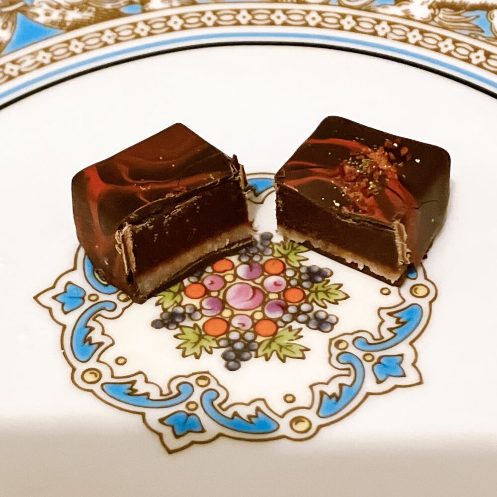 ウェッジウッド「フロレンティーン ターコイズ」の皿の上でカットされた、新作ボンボン「フランボワーズ」の断面図。表面に赤い流線模様が描かれたビターチョコレートの中に、フランボワーズ風味のガナッシュが隙間なく詰まっている様子が分かる。