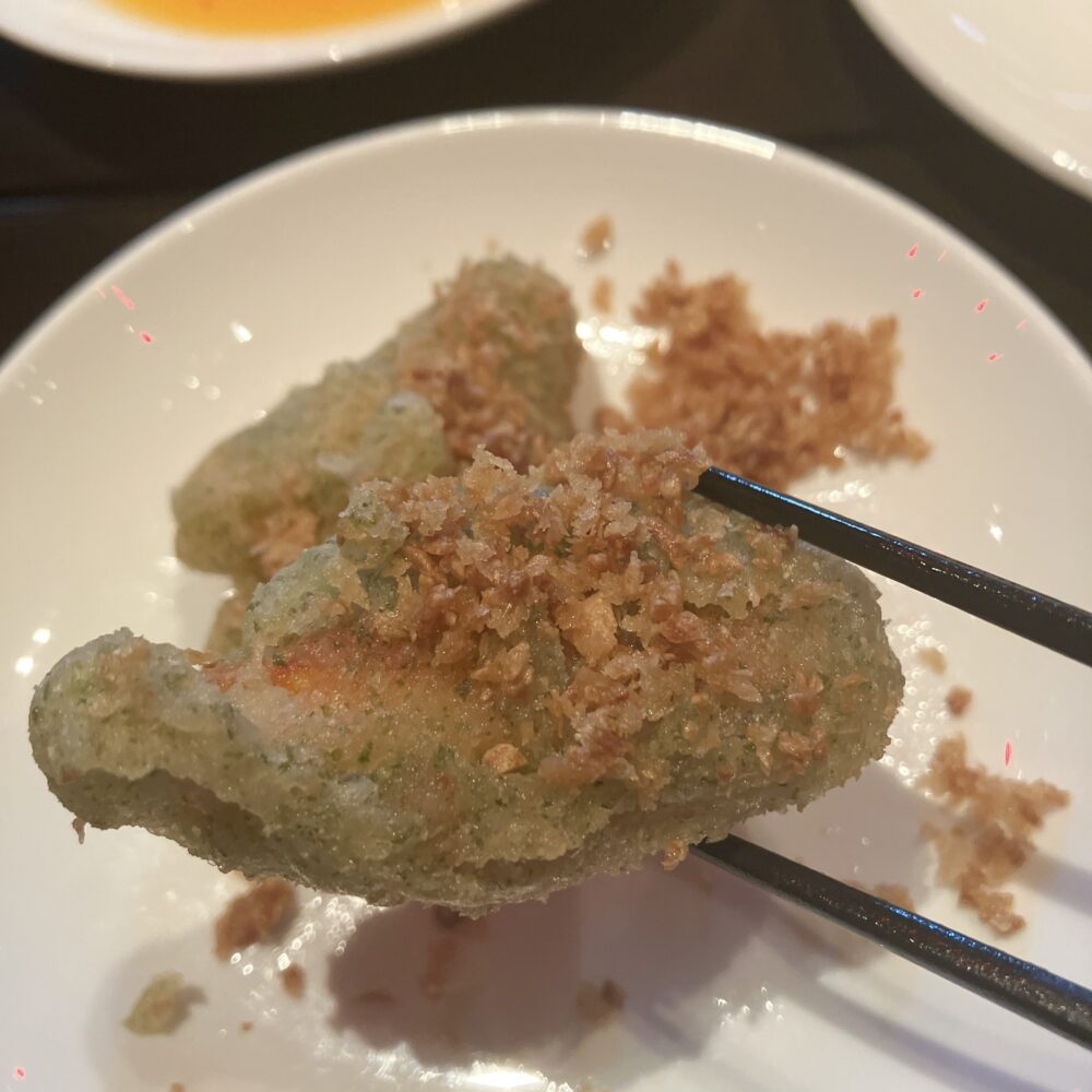 箸で持ち上げられた、緑色の衣が特徴的なズワイ蟹のフライドガーリック。香ばしいガーリックチップがまぶされている。