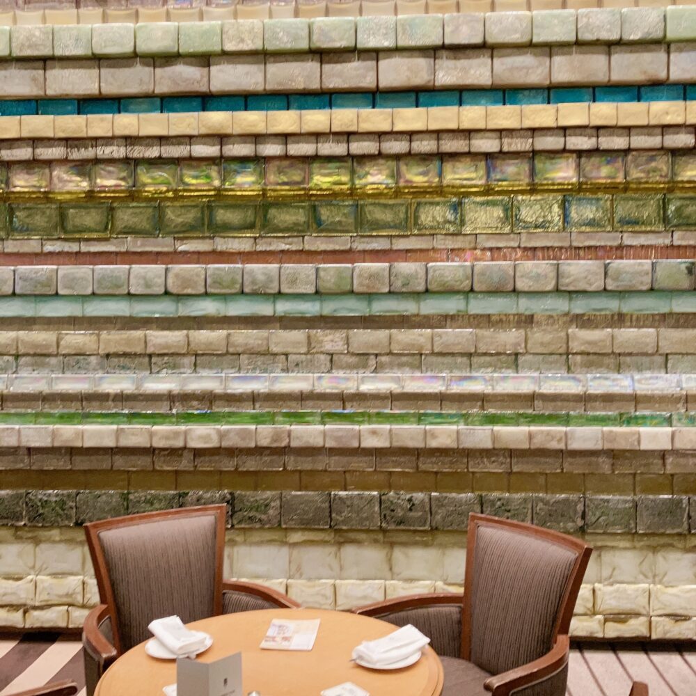 帝国ホテル「ランデブーラウンジ」にある多田美波作「光の壁」のクローズアップ写真。重厚な石のように見える様々な色や質感のガラスブロックが幾重にも積み重なっている。手前にはティータイム用の丸テーブルと2脚の椅子が置かれている。