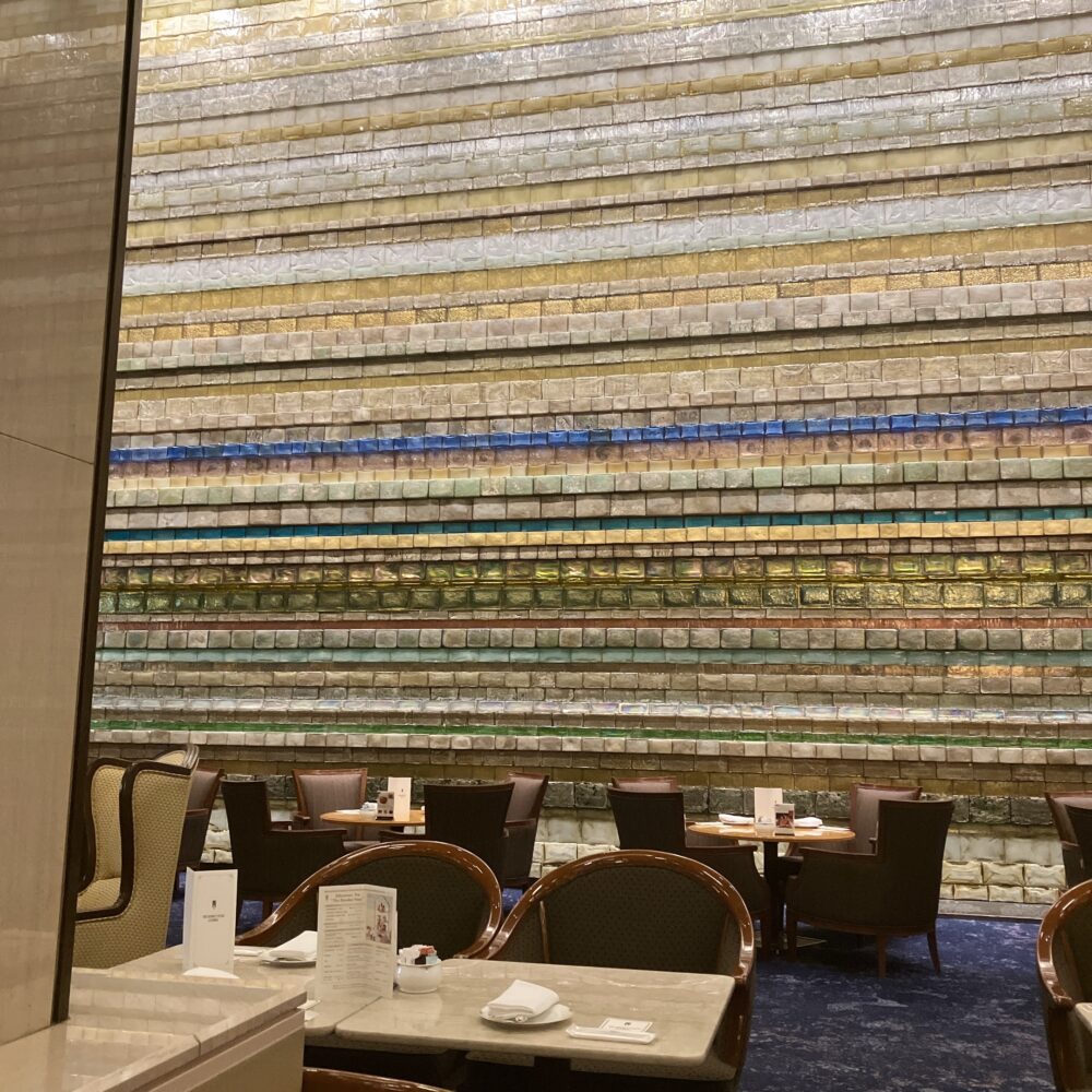 帝国ホテルのランデブーラウンジの壁面を覆う、多田美波作の「光の壁」。無数のガラスブロックが整然と積まれ、金や青、緑の光を放っている。手前にはアフタヌーンティーを楽しむためのテーブルとソファーが並んでいる。