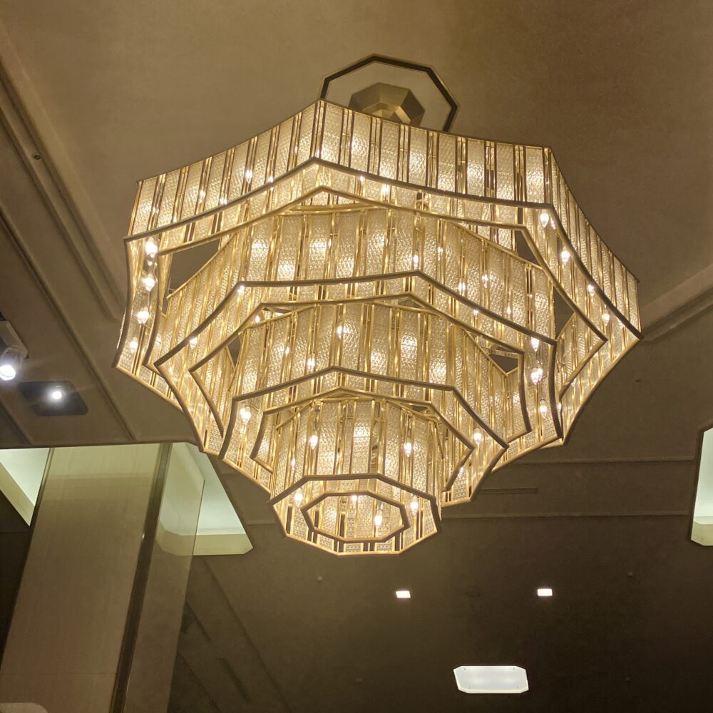 多田美波作の『薔薇のシャンデリア』を下から見上げた近距離写真。無数の四角いガラス板と真鍮のフレームが幾何学的に組み合わさり、全体として何層にも重なる薔薇の花びらを形成している。