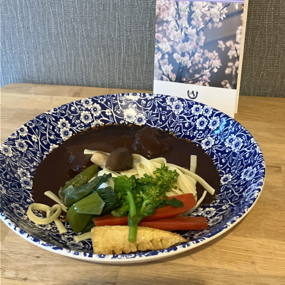 自宅の青と白の美しい花柄の皿に美しく盛り付けられた帝国ホテルのビーフシチュー。濃厚なブラウンのソースの上にはゴロゴロとした牛肉があり、手前にはヌードルと春らしい筍、菜の花、人参などが添えられている。奥には桜のポストカードが飾られている。