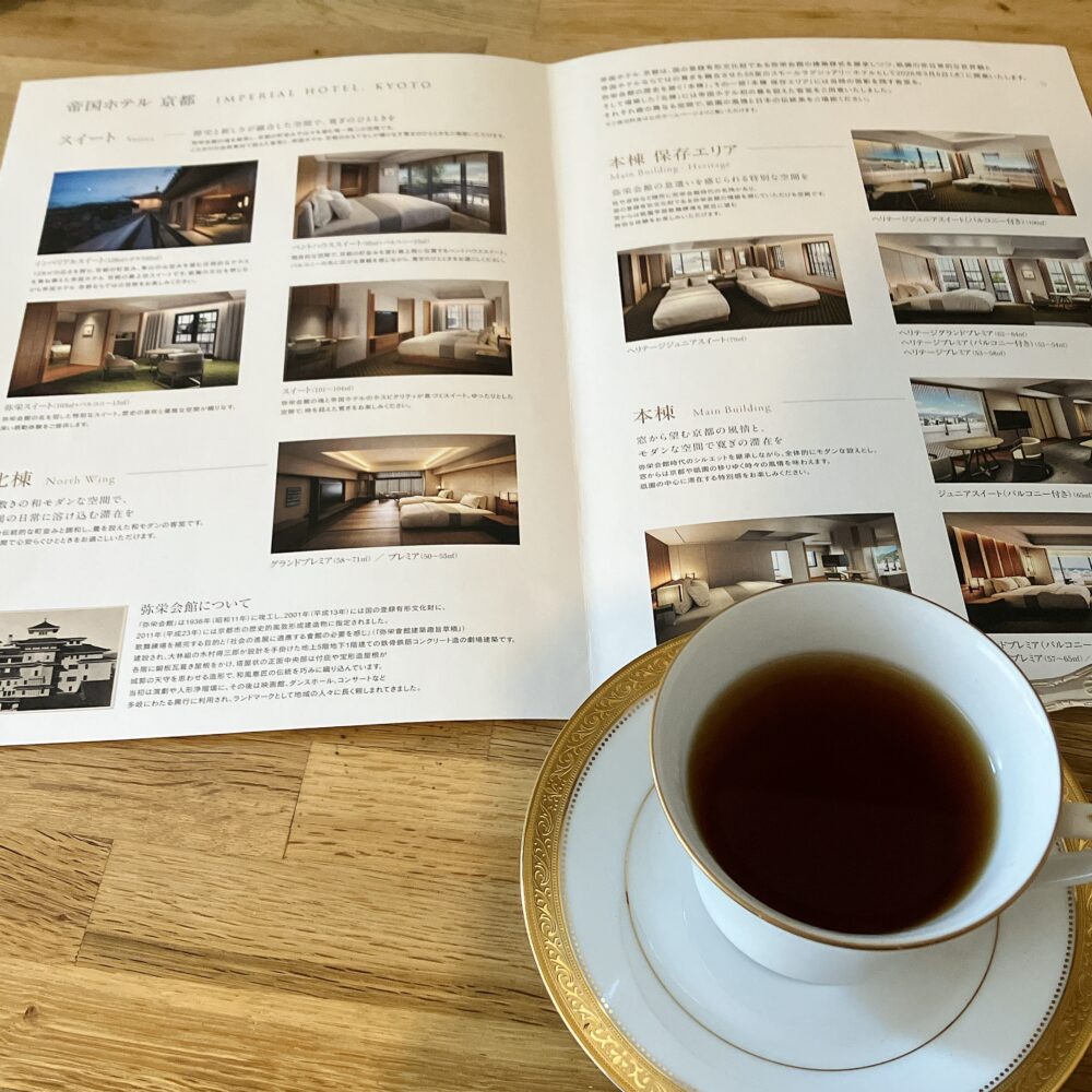 自宅のデスクで紅茶を楽しみながら、帝国ホテル 京都の客室パンフレットを広げている様子。「北棟」「本棟」などの客室写真が掲載されている。
キャプション： 予約日を前に、自宅でパンフレットを広げて作戦会議。この時間が一番ワクワクします。