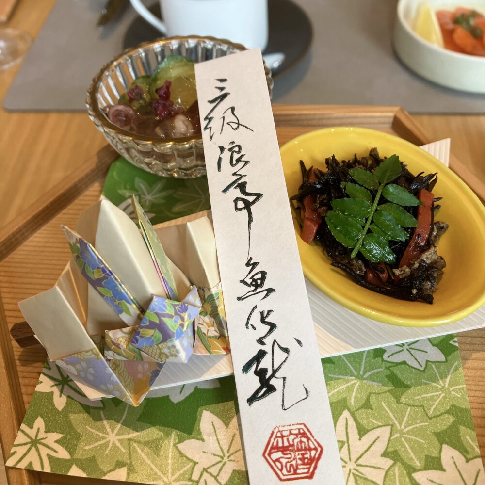 お重箱の蓋の上に盛り付けられた小鉢「ホタルイカのジュレ掛け」と「ひじきの煮物」。美しい折り紙の装飾が添えられている