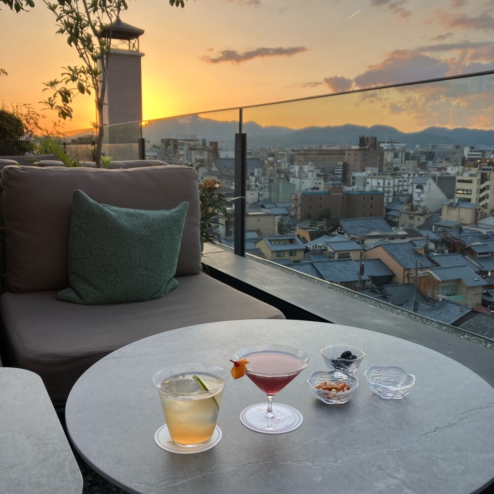 帝国ホテル京都のルーフトップバーのソファ席から見た、夕暮れの京都の街並みと東山の景色。テーブルには、2種類のドリンクとスナックが置かれている。
