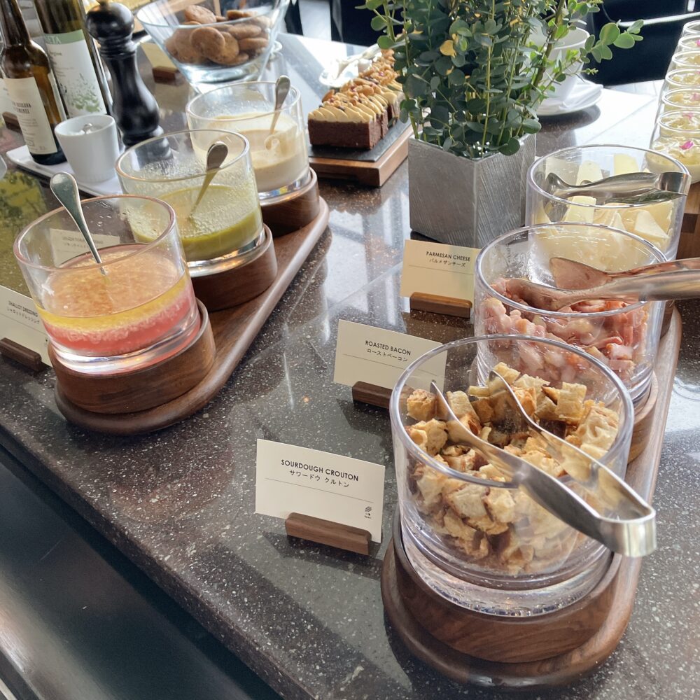 サラダ用のトッピングコーナー。美しくグラスに盛られたシャロットドレッシング、カリカリのローストベーコン、サワードウクルトン、削りたてのパルメザンチーズが並んでいる。