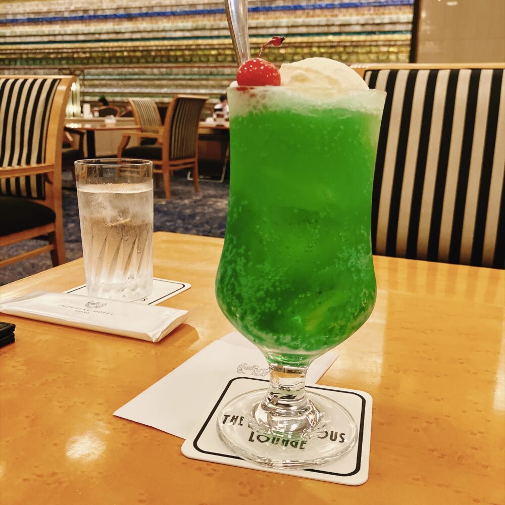 ランデブーラウンジのテーブルに置かれた色鮮やかなメロンソーダ。背景にはストライプ柄の椅子と象徴的なモザイク壁画「光の壁」が見える。