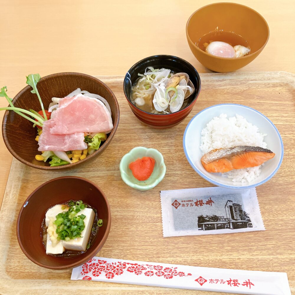 焼き鮭、ご飯、あさりの味噌汁、湯豆腐、生ハムサラダ、明太子、温泉卵が美しく盛り付けられた和食メインの朝食トレイ。
