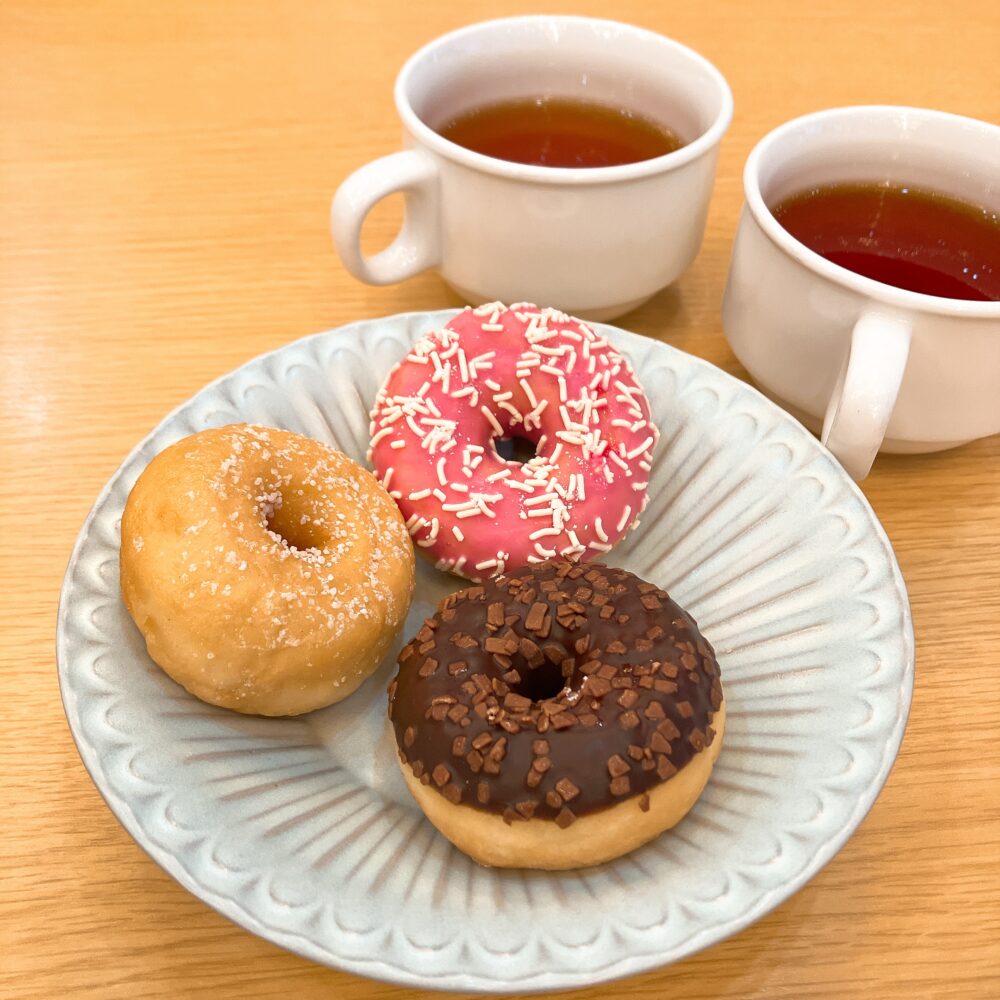 シュガー、ストロベリー、チョコレートの3種類のドーナツがお皿に乗っており、奥には紅茶のカップが2つ並んでいる。
