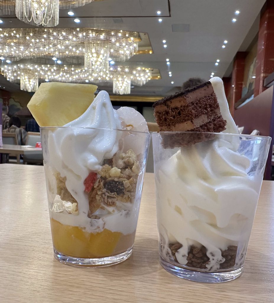 ホテル櫻井の朝食バイキングで、ソフトクリームにフルーツやシリアル、チョコレートケーキをトッピングして作った2種類のオリジナルパフェ。背景には豪華なシャンデリア。