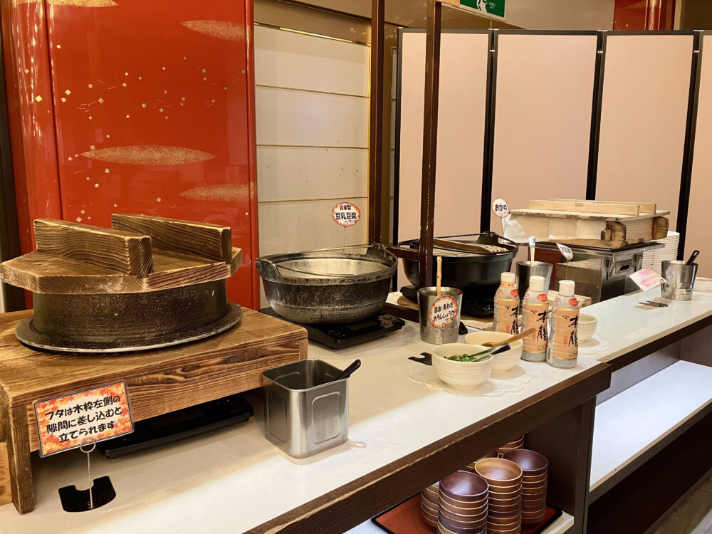 「自家製豆乳豆腐」と「おかゆ」が並ぶ和食コーナー。大きな鍋や木製の蓋が使われており、手前には薬味や醤油が置かれている。